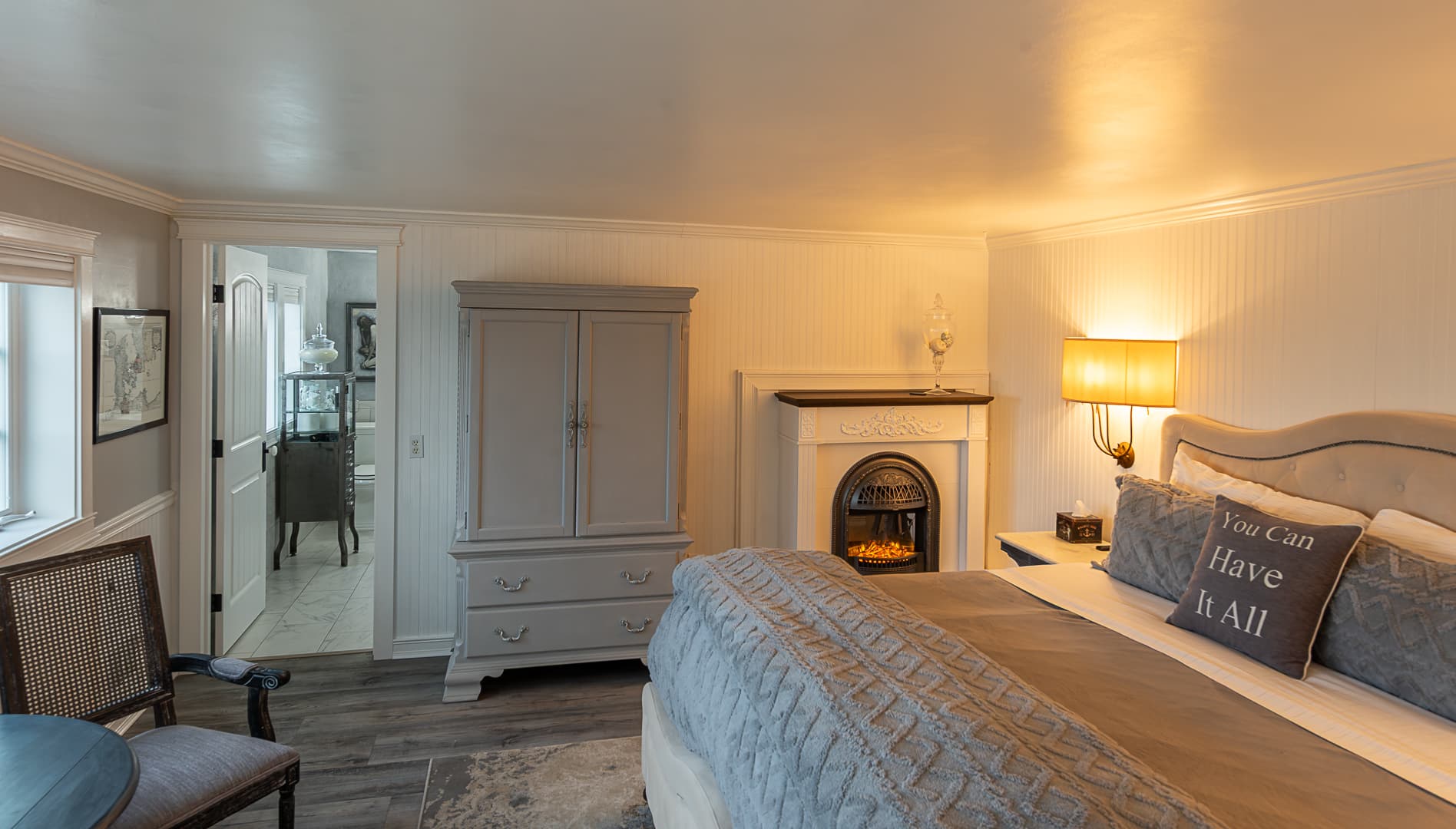 A cozy bedroom suite featuring a king-size bed with grey patterned linens, a warm glowing fireplace, and a classic grey armoire, with an open doorway leading to a bright adjacent room.