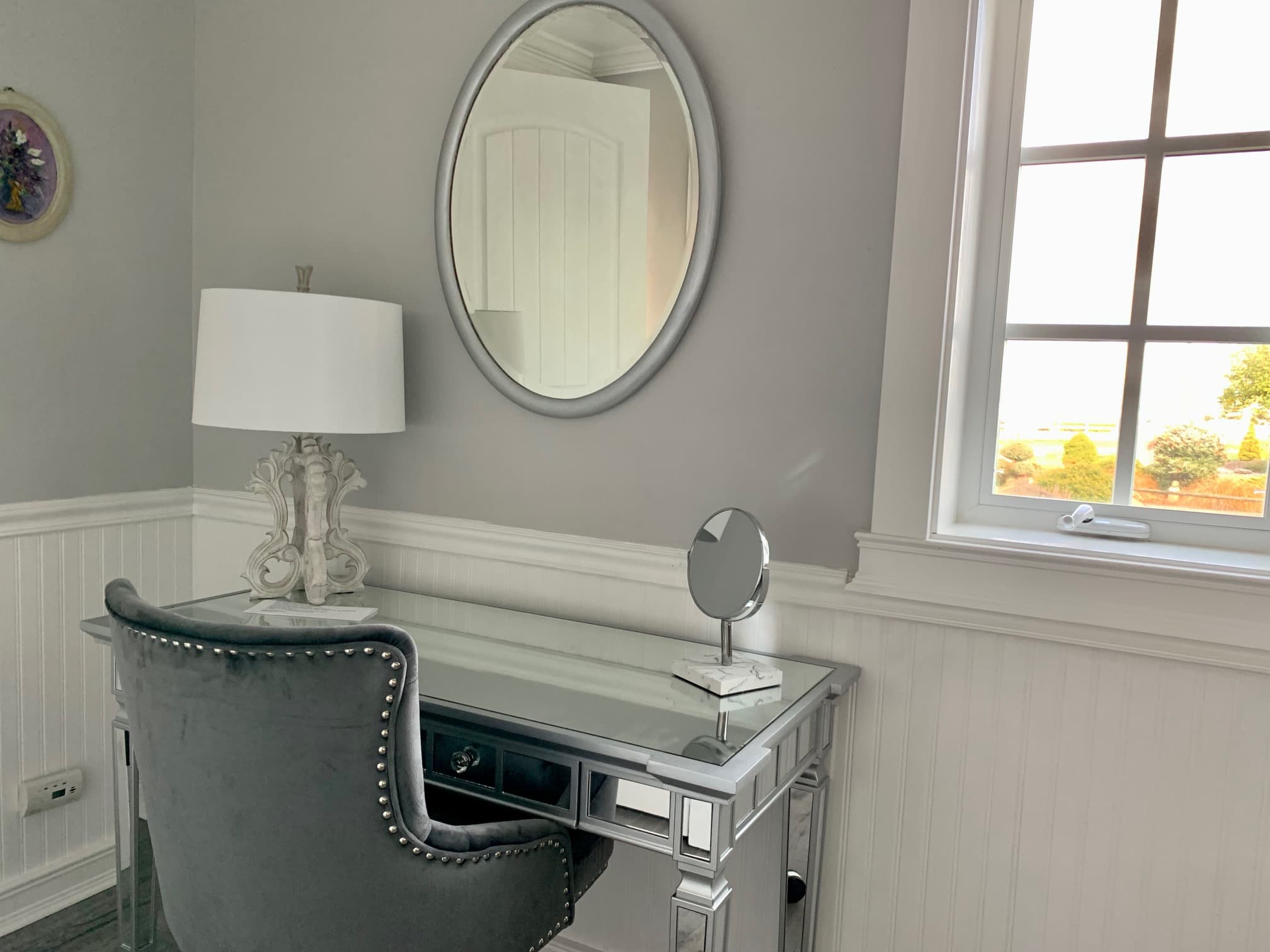 A sophisticated vanity area featuring a mirrored desk with silver trim, a tufted grey velvet chair, and an oval silver-framed mirror mounted on a grey wall above white wainscoting.