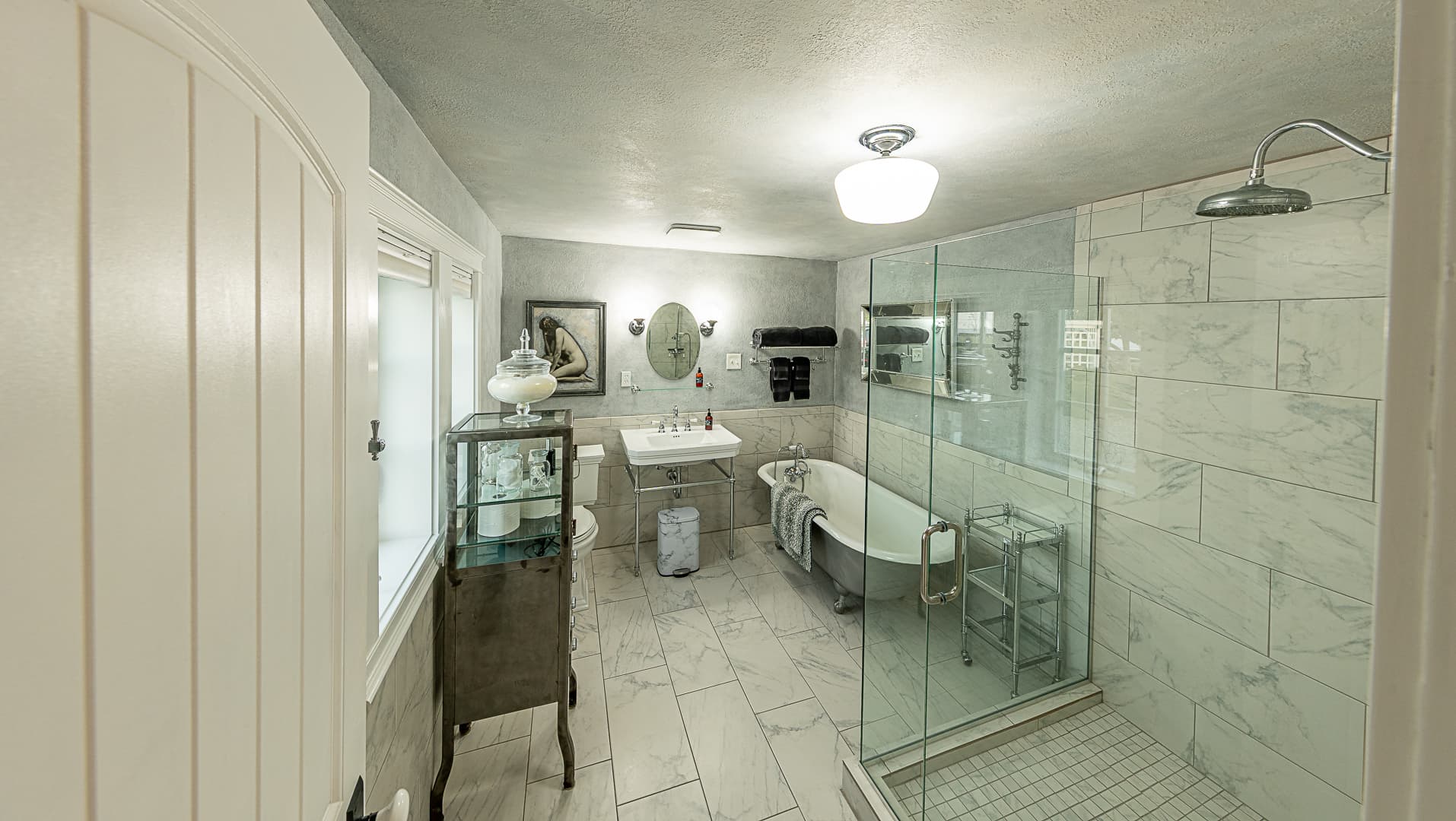 A spacious, luxury bathroom with white marble-style tiling, featuring a large glass-enclosed walk-in shower on the right and a classic clawfoot soaking tub.