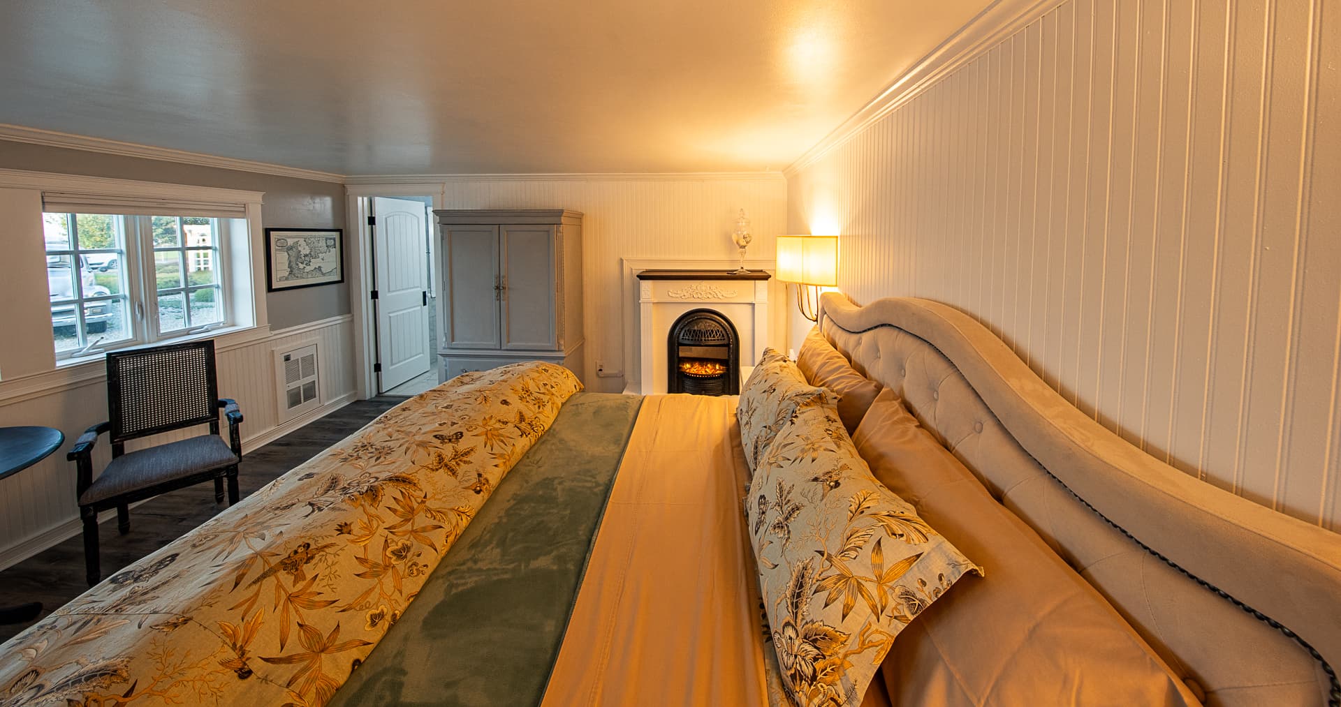 An wide-angle view of a cozy bedroom suite featuring a king-size bed with seashell-patterned pillows, a warm fireplace, and a grey armoire, with white beadboard wainscoting along the walls.