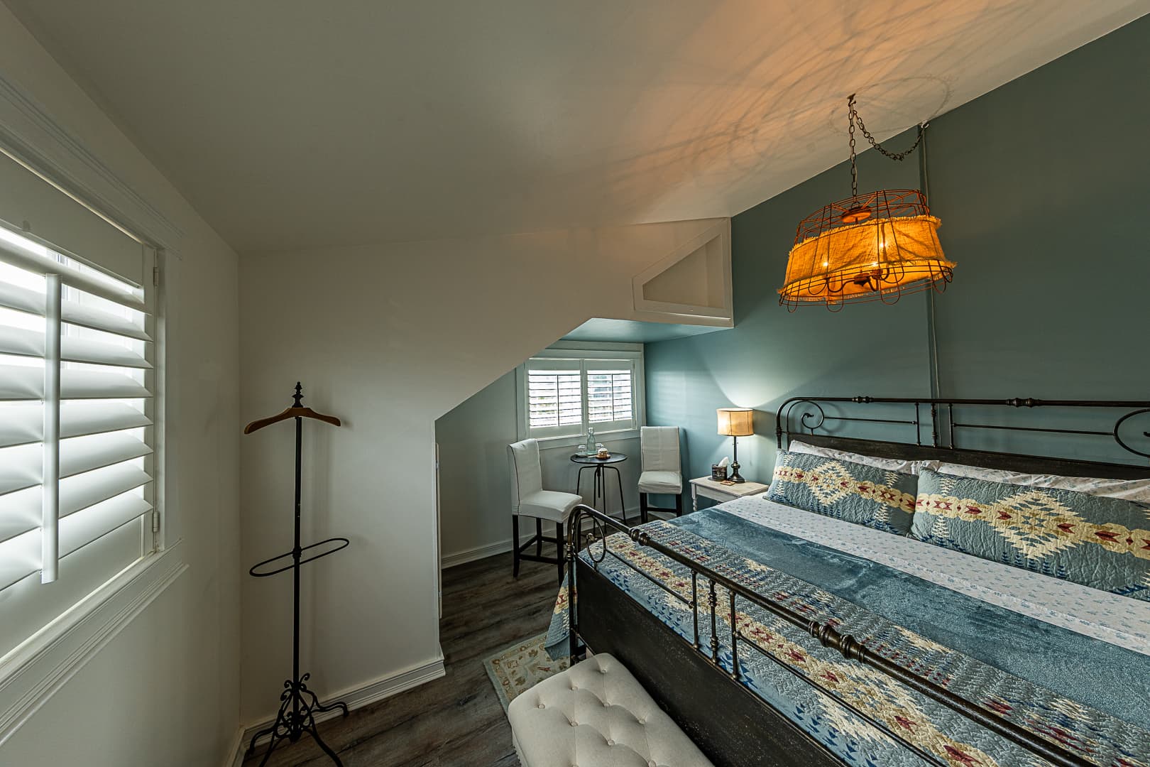 A bedroom suite featuring a king-size bed with blue southwestern-patterned linens, a unique orange-fringe pendant light, and a cozy seating nook by a shuttered window, all under a sloped ceiling.