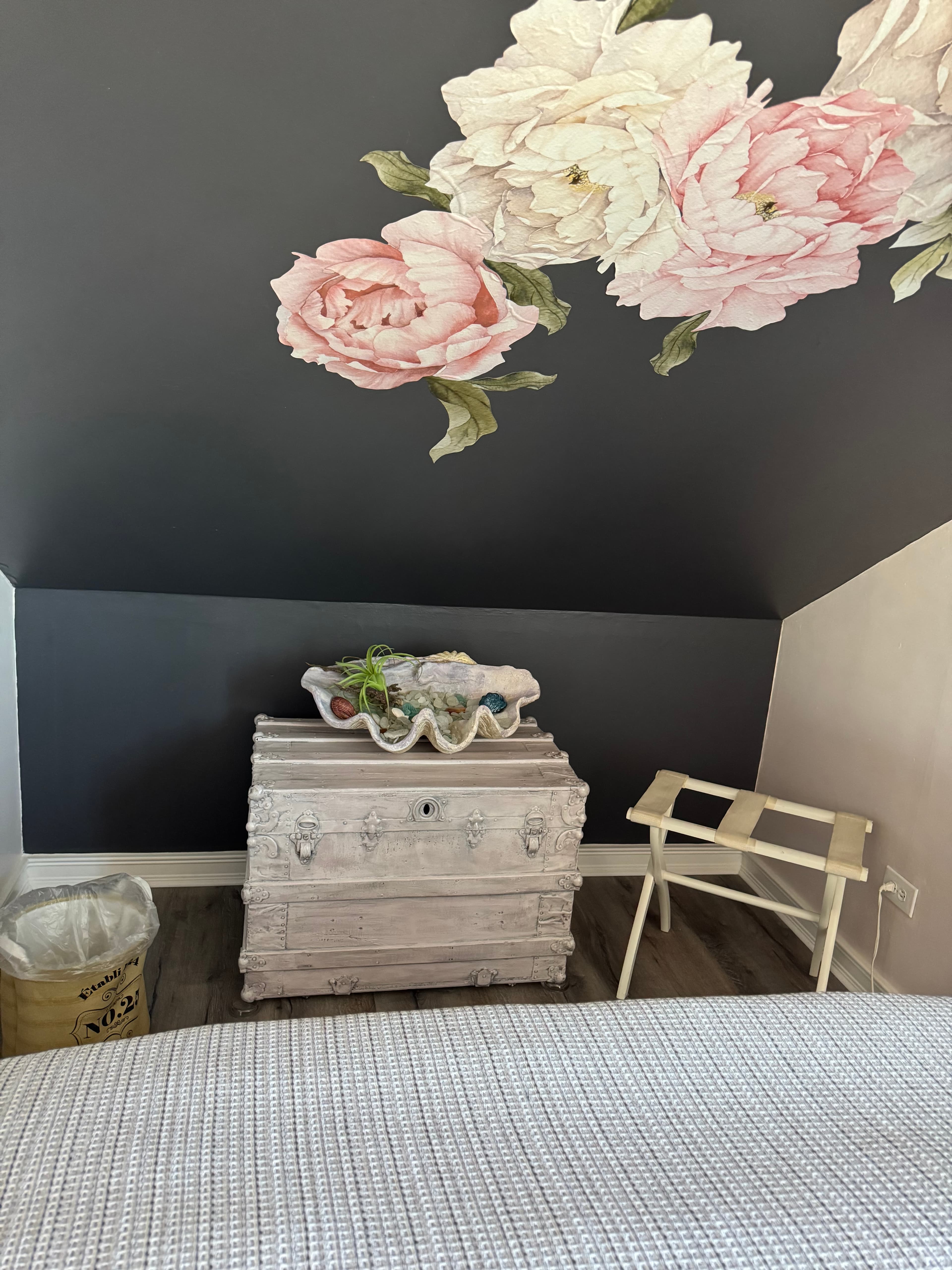 An attic-style bedroom nook featuring a grey slanted wall adorned with large pink and white floral decals above a vintage white wooden trunk and a folding luggage rack.