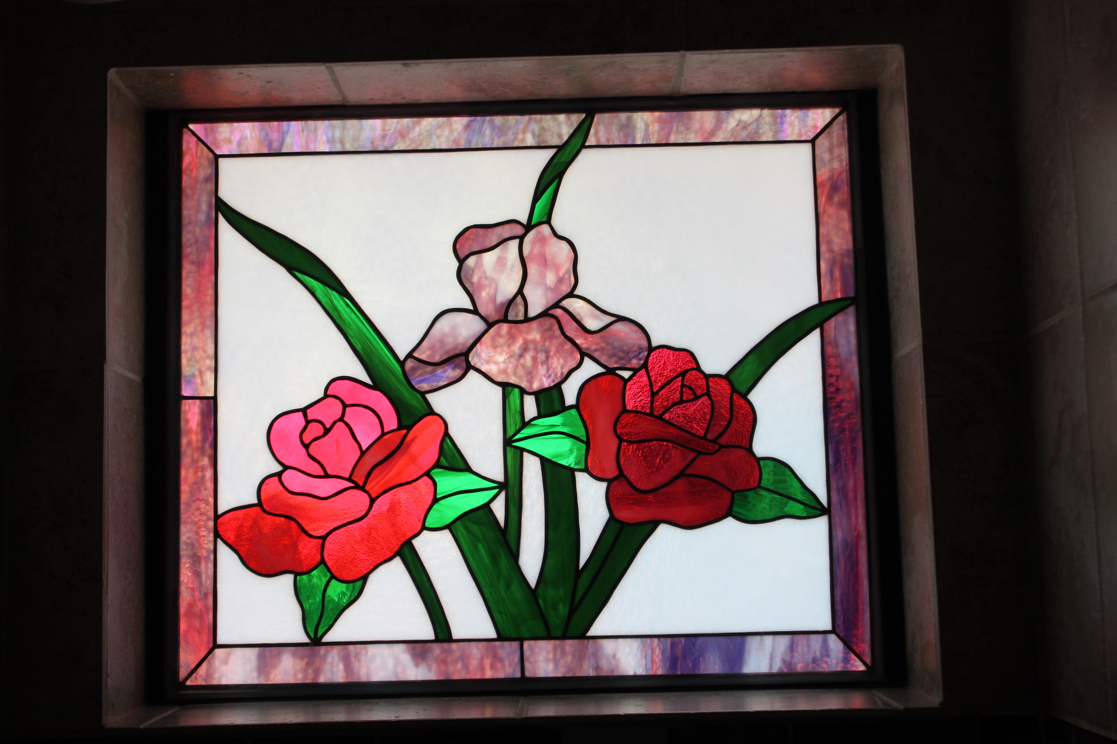 A close-up of a square stained-glass window featuring vibrant pink and red roses with green leaves against a white background, framed by a purple marbled border.
