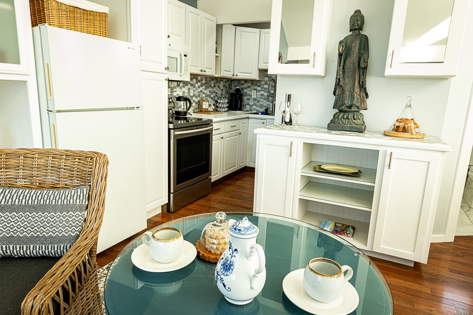 A bright studio kitchenette featuring white cabinetry, a full-size refrigerator, and a small range, viewed from a seating area with a wicker chair and a glass-top table set for coffee.