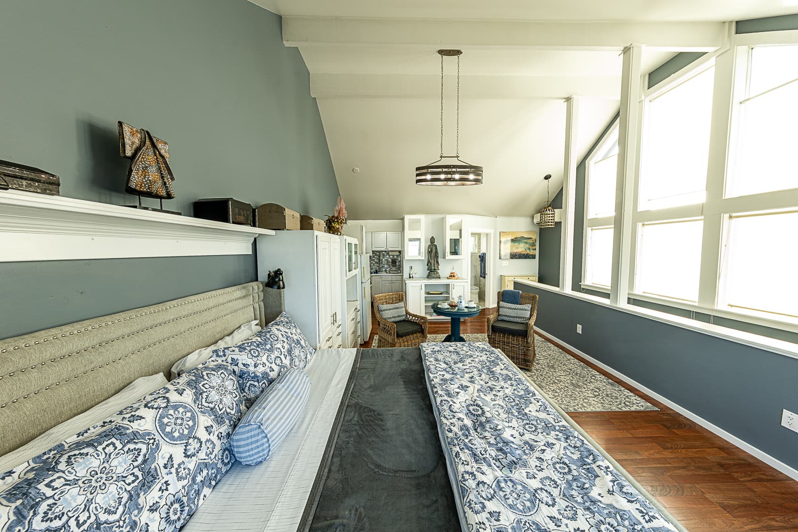 A wide view of a modern studio guest suite featuring a large bed with blue patterned linens in the foreground, floor-to-ceiling windows on the right, and a kitchenette with a dining nook in the background under a vaulted ceiling.
