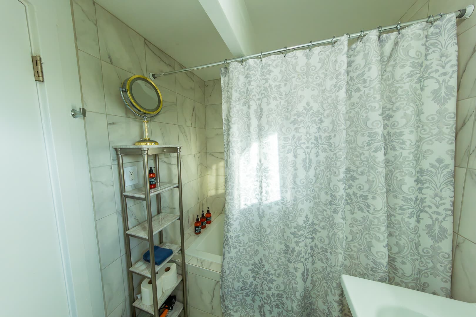 An elegantly tiled bathroom featuring a soaking tub/shower enclosed by a grey and white patterned shower curtain, complemented by marble-style wall tiles and a silver shelving unit holding toiletries and a vanity mirror.