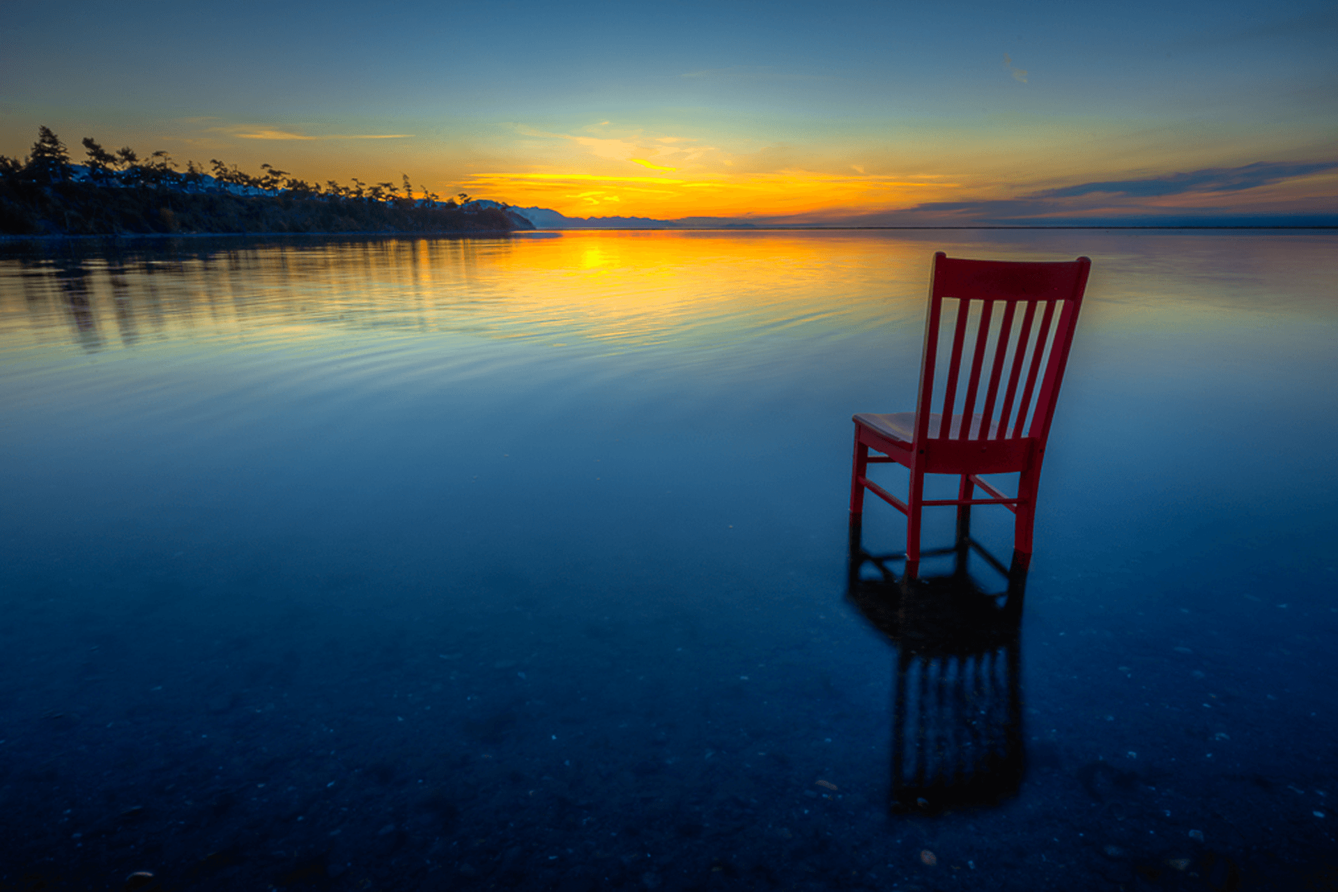 A red chair stands in calm water at sunset, reflecting the colorful sky.