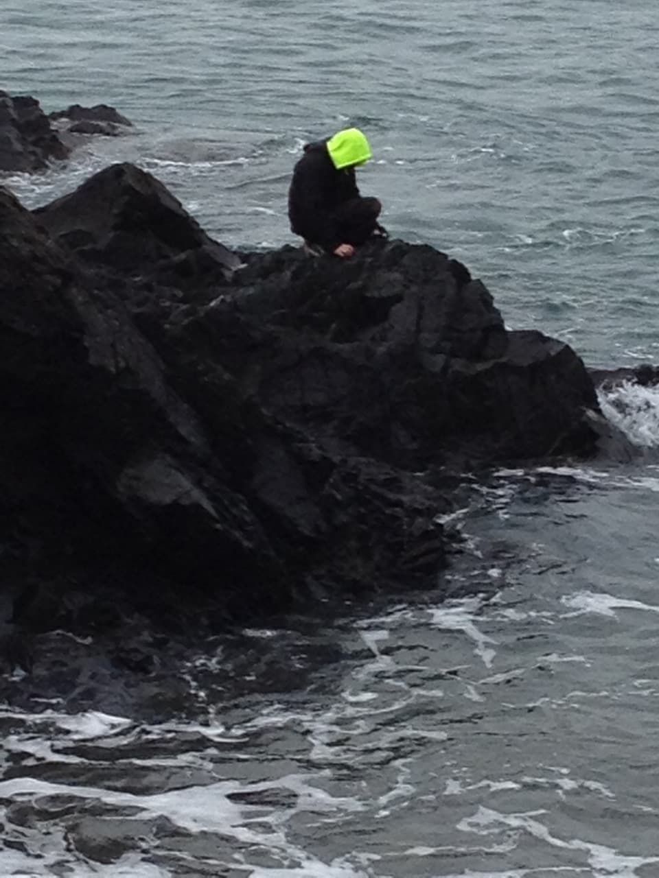 A person wearing a bright yellow hat sits on a rock by the shoreline, surrounded by water.