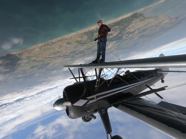A person stands on the wing of a biplane flying upside down over a landscape.