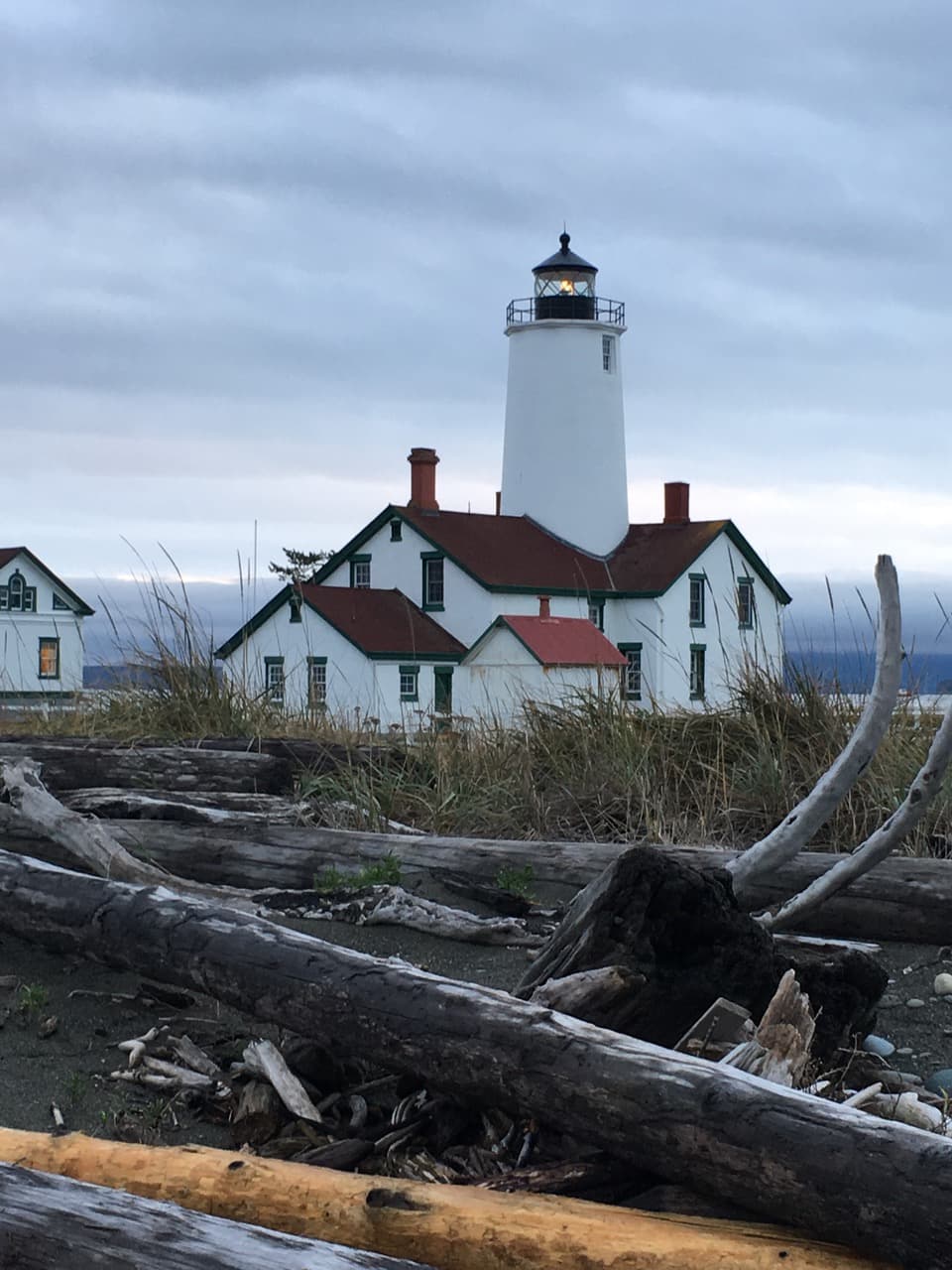 A white lighthouse with a black lantern stands next to a historical building amidst driftwood on a cloudy beach.