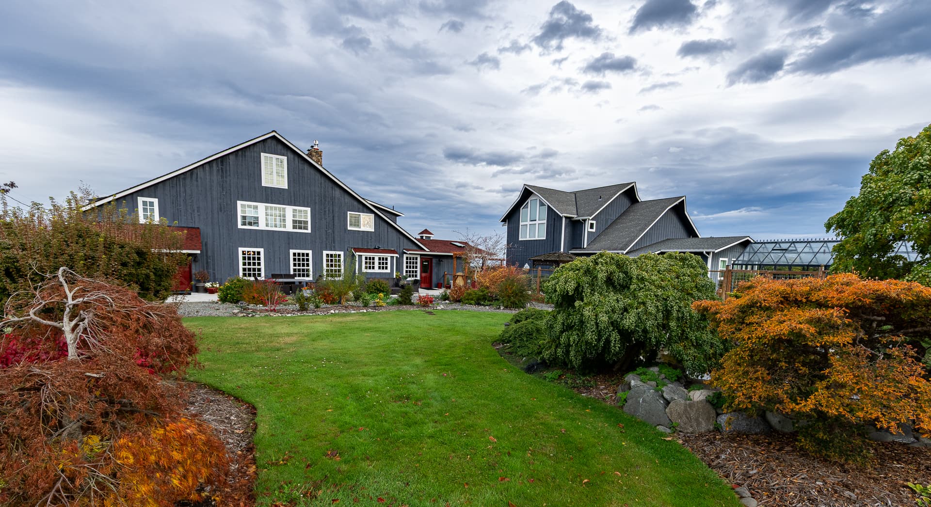 A large, dark-colored house surrounded by lush green grass and colorful autumn foliage under a cloudy sky.