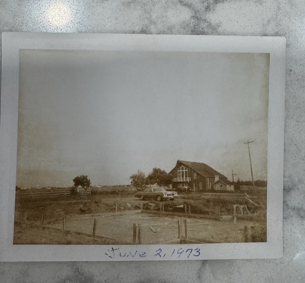 A sepia-toned photograph from June 2, 1973, showing a house, a truck, and an empty lot surrounded by farmland.