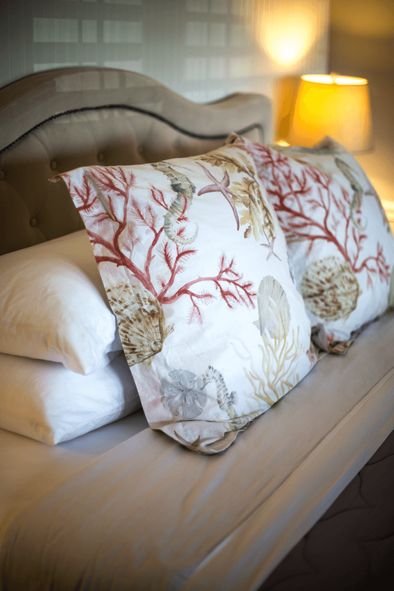A close-up of decorative pillows with coral and shell patterns on a neatly made bed beside a lamp.
