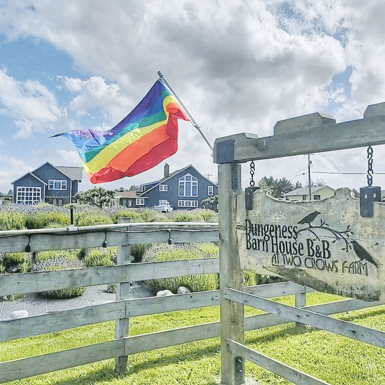 A rainbow flag waves next to the sign for Bungeness Barn House B&B at Two Crows Farm, amidst a scenic rural backdrop.