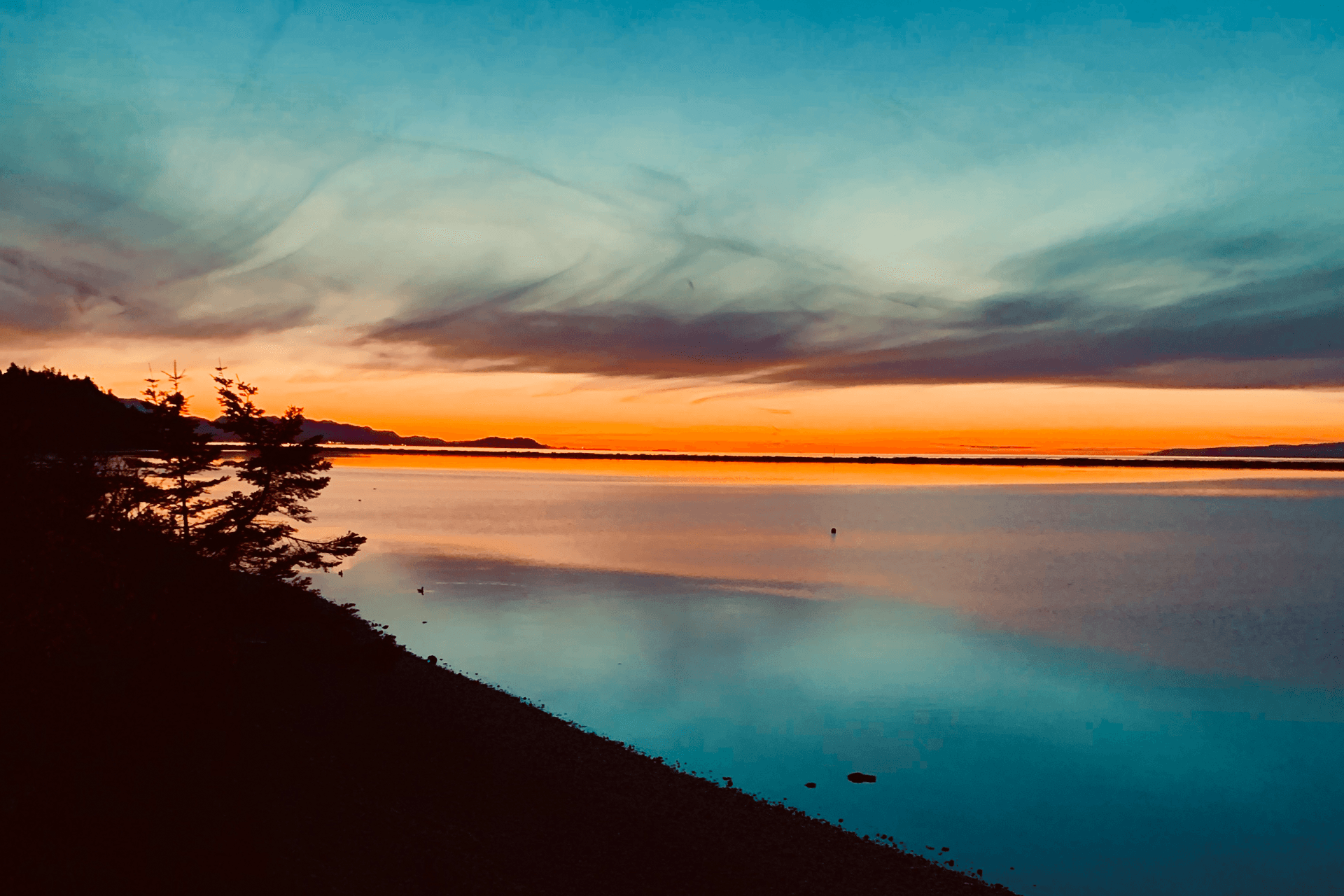 A serene sunset reflecting vibrant colors over a calm body of water, framed by silhouettes of trees.