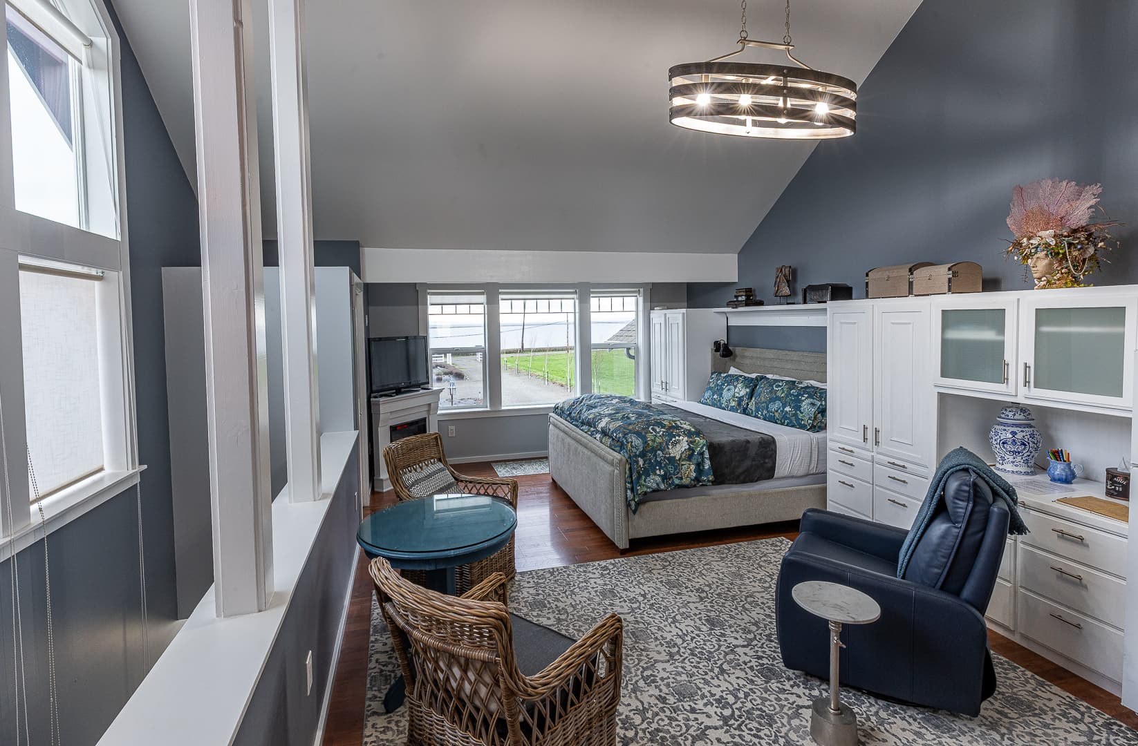 A bright studio kitchenette featuring white cabinetry, a full-size refrigerator, and a small range, viewed from a seating area with a wicker chair and a glass-top table set for coffee.