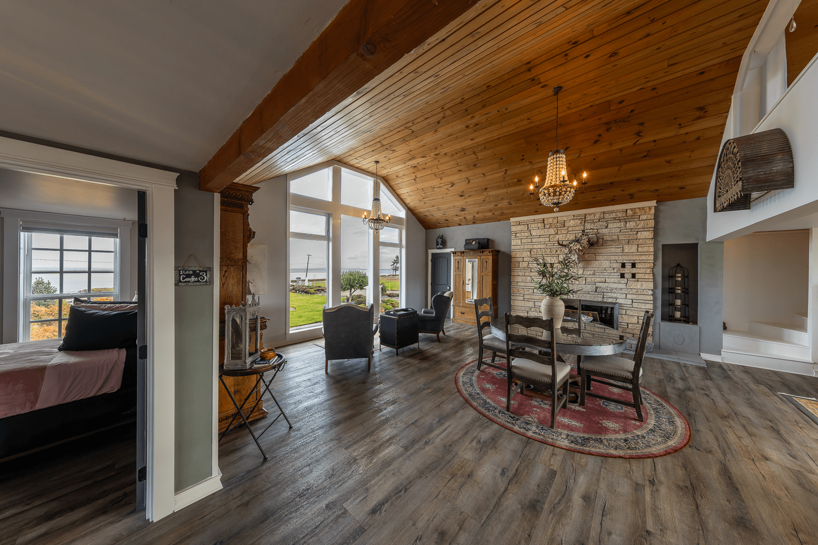 A bright, spacious living area featuring wooden beams, a dining table, and large windows overlooking greenery.