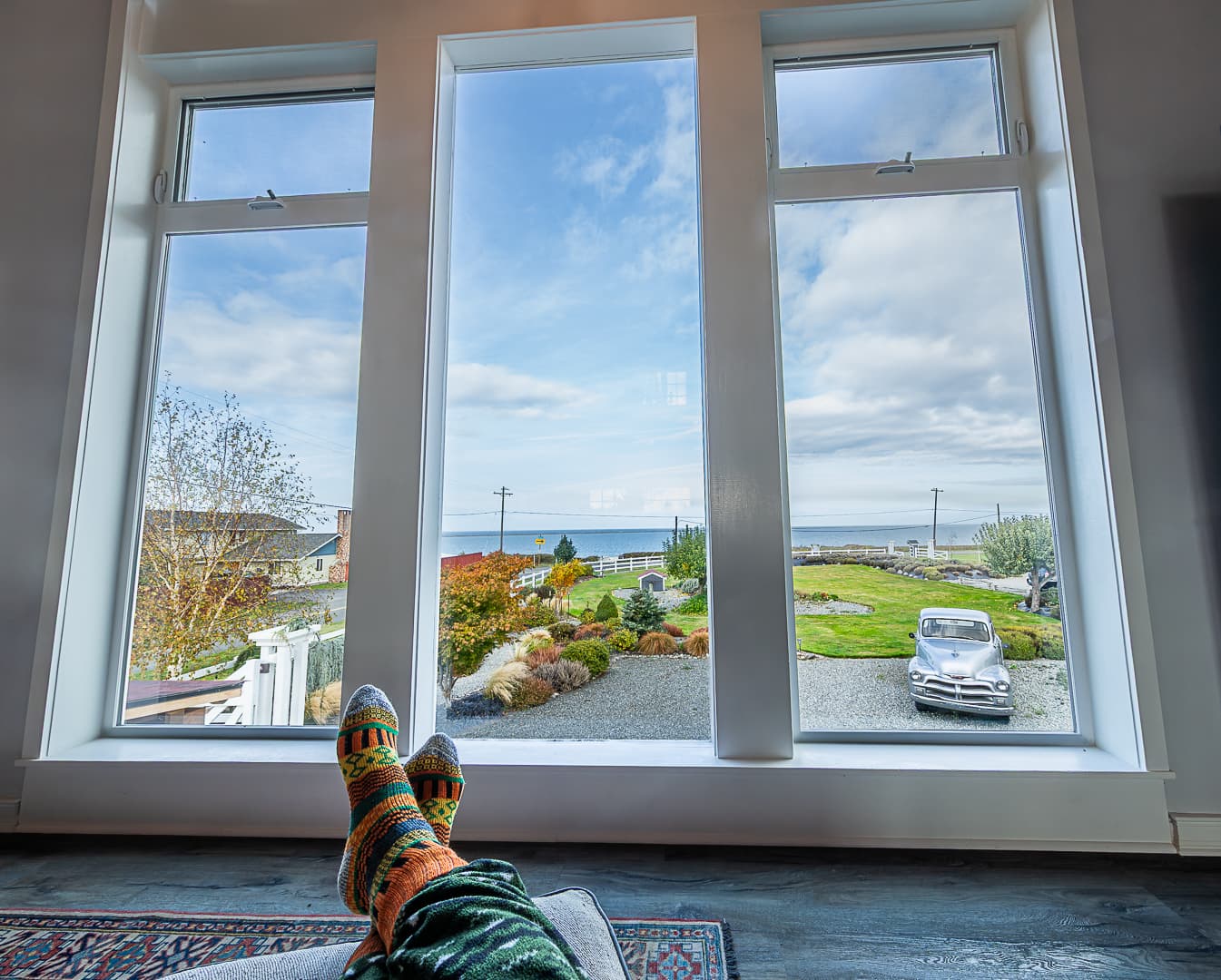 A person relaxes indoors, feet propped up, gazing out through large windows at a scenic view of the ocean and garden.